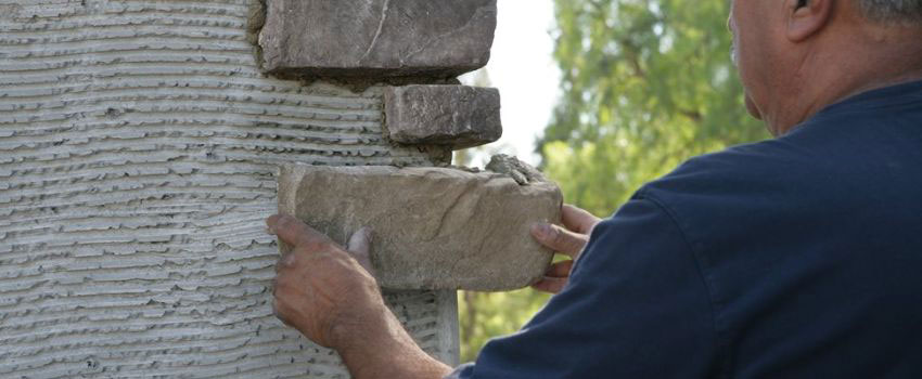 A worker applying a scratch coat onto a brick wall in preparation for cultured stone installation in Edmonton, AB. The mortar is spread evenly with a trowel, creating a rough texture for the cultured stone to adhere to securely.