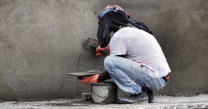 A close-up view of a professional applying traditional cement parging on the exterior foundation wall of a home in Edmonton, AB. The trowel spreads a smooth, even layer of the cement mixture to create a protective coating over the masonry surface.